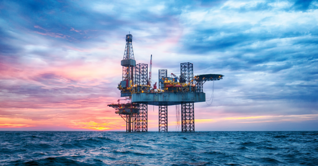 Offshore oil platform standing on stilts in the sea at sunset, lit by lights and cranes visible against a colorful sky.